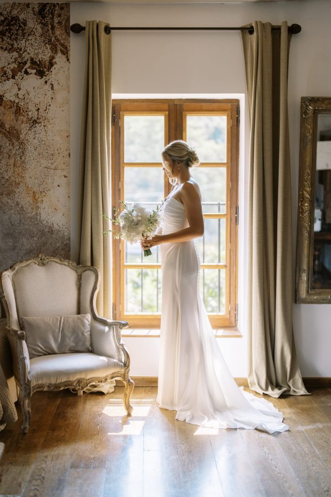Mariée en robe blanche tenant un bouquet, debout face à une fenêtre dans une pièce élégante et lumineuse.