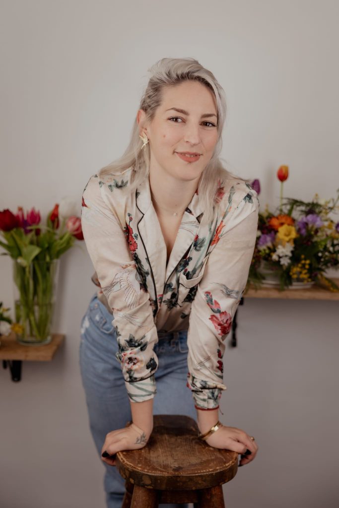 Portrait d’une femme souriante debout derrière un tabouret, devant des fleurs colorées dans un atelier