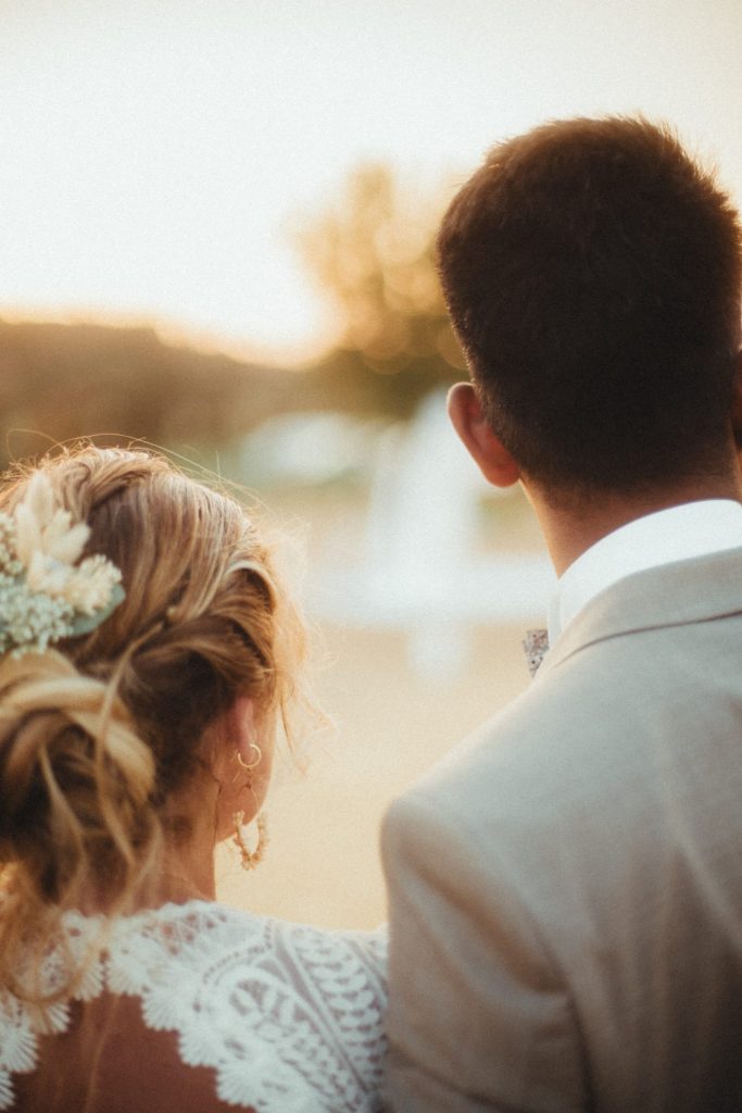 Couple de mariés vu de dos au coucher du soleil, la mariée portant une coiffure fleurie et une robe en dentelle.