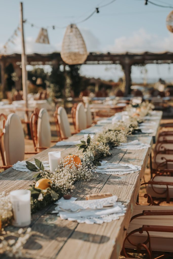 Grande table de réception de mariage en extérieur, décorée de fleurs, feuillages, bougies et agrumes, avec des chaises alignées sous un ciel ensoleillé.